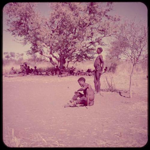 Dance: /Ti!kay sitting and putting on his dance rattles, Gau standing beside him, with people sitting under a tree in the background