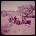 Dance: Men and two little boys dancing in a line through a circle of women sitting, with people sitting under a tree in the background, view from an expedition truck