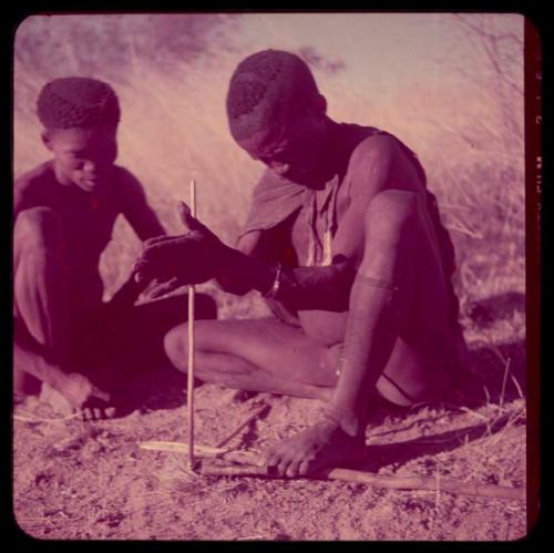 Fire-Making: ≠Gao (headman of Band 3) sitting with his brother, "/Qui Hunter," using firesticks and grass tinder to make a fire