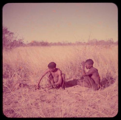 Fire-Making: ≠Gao (headman of Band 3) sitting with his brother, "/Qui Hunter," using firesticks and grass tinder to make a fire, distant view