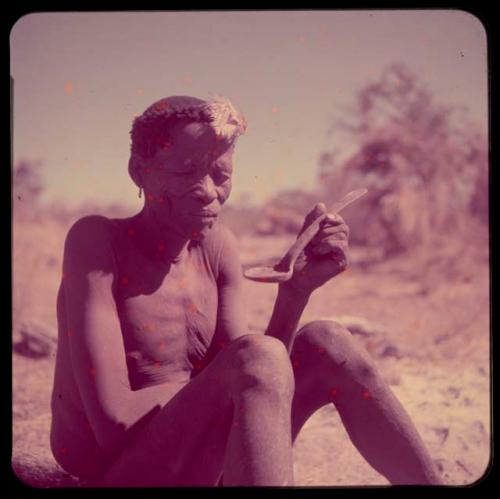 Food, Eating: "Old !Naishi" (headman of Band 5) wearing an ostrich feather in his hair, sitting and holding a spoon