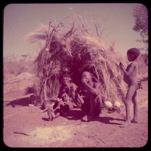 Food, Eating:  //Kushay and "/Qui Navel" sitting in their skerm, eating, with one of their sons standing next to them