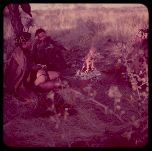 Food Gathering, Tsi: Khuan//a of Band 4 (wife of //Ao) wearing copper rings, sitting with another woman (possibly "Gao Medicine's" sister) next to a fire on a gathering trip