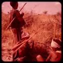 Two women sitting and digging to gather food, with another woman standing nearby