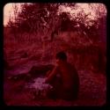 Man sitting by fire, with a bedroll on the other side, seen from behind