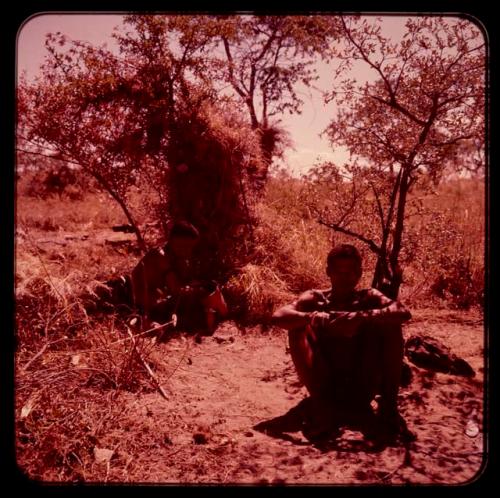 Man sitting on the ground, with a woman sitting under a leafy shelter in the background