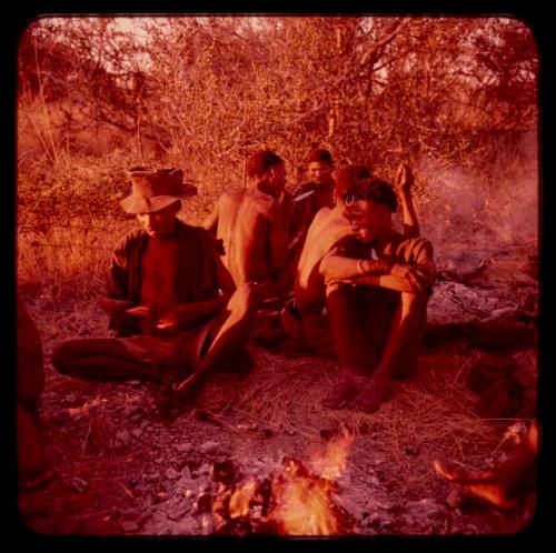 Group of men sitting near fire, including //Nami wearing a hat and pants