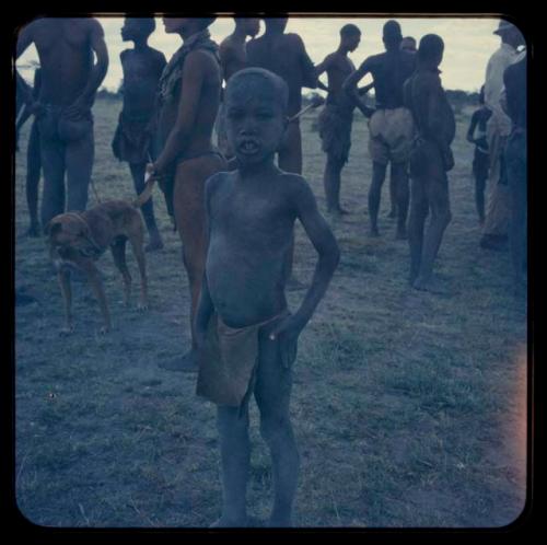 Boy standing, with a group of people and a dog in the background