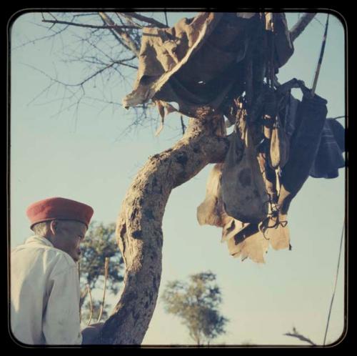 "Crooked /Qui" wearing Western clothes and standing beside a tree hung with gear