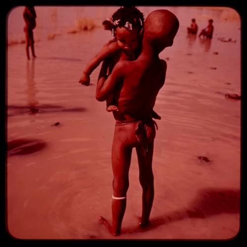 Boy standing in the water and holding a baby, seen from behind