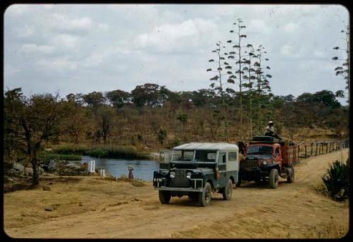 Two expedition trucks parked on the road to Cuchi, with a woman standing on the bank of a stream in the background