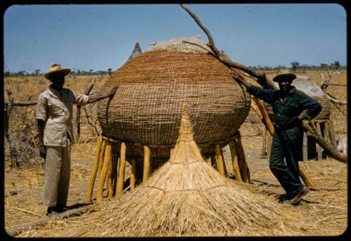 Elia and Philip Hameva standing next to a large basket mounted on poles, with a thatched roof for it on the ground next to it