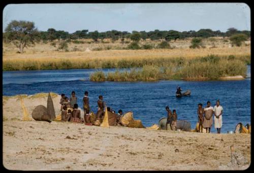 People standing and sitting along the Okavango River, with fish traps on the ground next to them