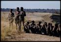 Men standing and sitting, waiting to be paid by Mr. Kruger for work on a fire break, with fire break in the background