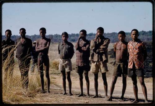 Men standing in line, waiting to be paid by Mr. Kruger for work on a fire break, one wearing traditional clothing, others wearing western clothing