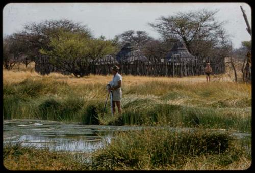 Eberhard von Koenen standing with camera equipment next to a waterhole, with a kraal in the background