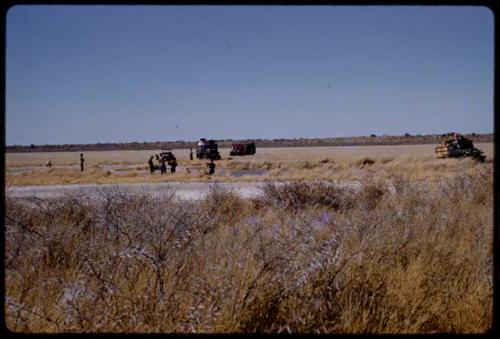 Expedition members and trucks by a waterhole, in the distance