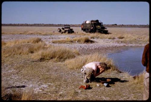 Expedition members and trucks by a waterhole, John Marshall washing his face