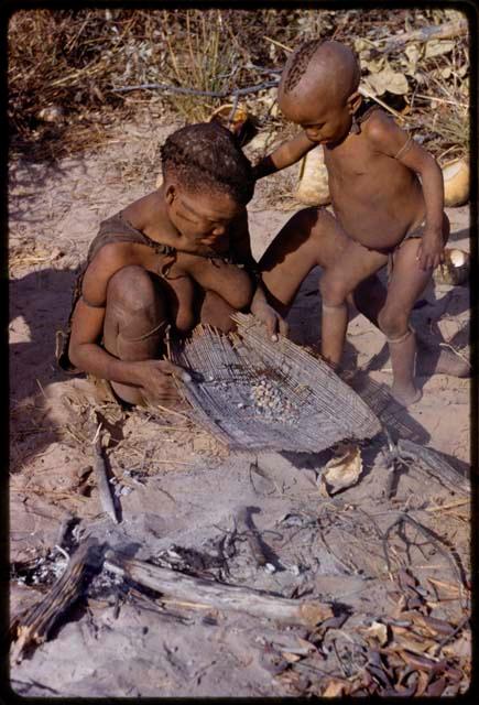 Tsekue roasting tsama melon seeds, N!whakwe standing next to her