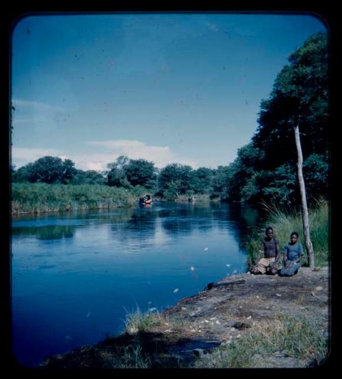 Man and a woman kneeling down on the bank of the Okavango River, with a barge from a distance