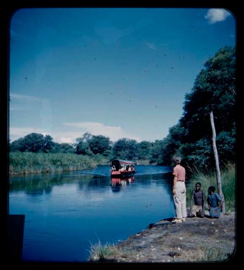 Man and a woman kneeling down and Lorna Marshall standing next to them on the bank of the Okavango River, with a barge from a distance