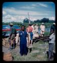 Group of people including the expedition members on the bank of the Okavango River; a man is holding fish with both his hands in the foreground and a barge is shown in the background