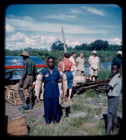 Group of people including the expedition members on the bank of the Kavango River; a man is holding fish with both his hands in the foreground and a barge is shown in the background