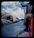 Man poling a barge toward a rocky shore of the Okavango River, seen from behind