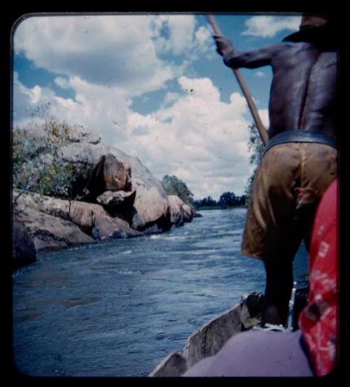 Man poling a barge toward a rocky shore of the Kavango River, seen from behind