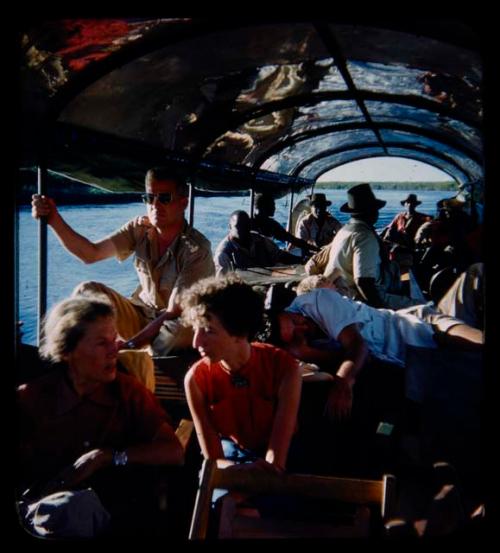 Group of people riding in a barge on the Kavango River, including expedition members, Anneliese Scherz, Lorna Marshall, and C.J. Mathias