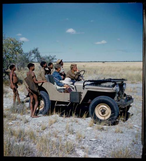 Expedition: Men standing behind the expedition Jeep, one carrying hunting equipment, with John Marshall in the driver's seat and Lorna Marshall sitting next to him