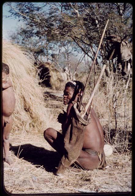 Carrying: Man holding a bag containing a bow and long sticks on his shoulder, sitting next to a skerm, with a child standing next to him