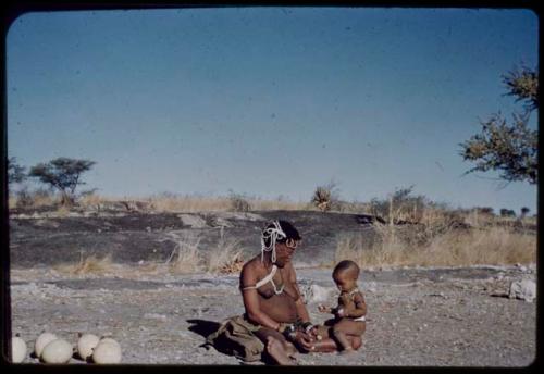 Children, Eating: !U sitting by the pan holding !Ungka Norna (after her haircut) on her lap, with ostrich egg shells beside her