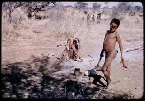 Children, Eating: Tsamgao (son of ≠Toma and !U) standing and holding a knife, with !U, !Ungka Norna and an unidentified person cooking in ashes near him