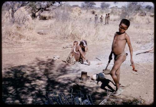 Children, Eating: Tsamgao (son of ≠Toma and !U) standing and holding a knife, with !U, !Ungka Norna and an unidentified person cooking in ashes near him