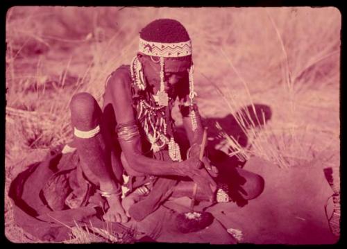 /Gam wearing a headdress of Western beads, making a bead by drilling a hole in a broken piece of ostrich eggshell