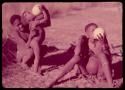 /Gunda (!Nai's husband) helping "Little ≠Gao" to drink from an ostrich eggshell, with a woman sitting next to them also drinking from an eggshell