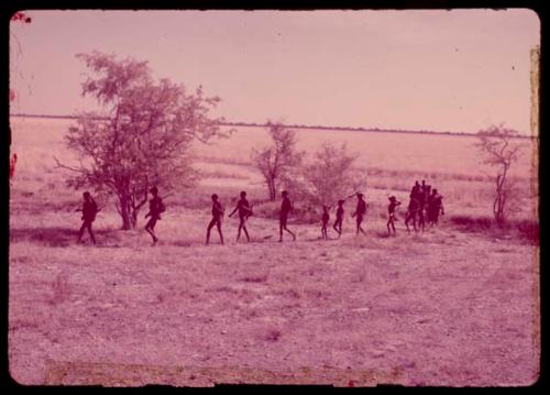 People walking in a line through the grass at the edge of a dry pan, with the men walking at the front of the line