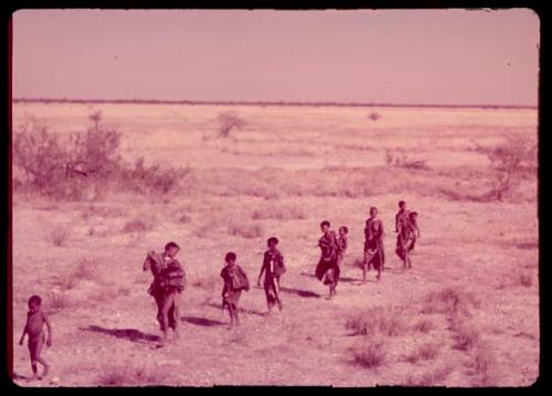 People walking in a line through the grass at the edge of a dry pan, with the women walking behind the men