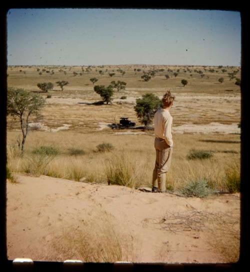 John Marshall standing, view from behind, with an expedition truck parked under a tree in the background