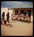 Group of children standing in front of a school, with three men standing and sitting in front of them