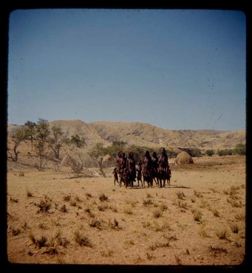 Group of women and children standing, with huts in the background, distant view