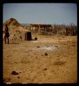 Person standing next to a hut, with a corn storage crib and a fence in the background