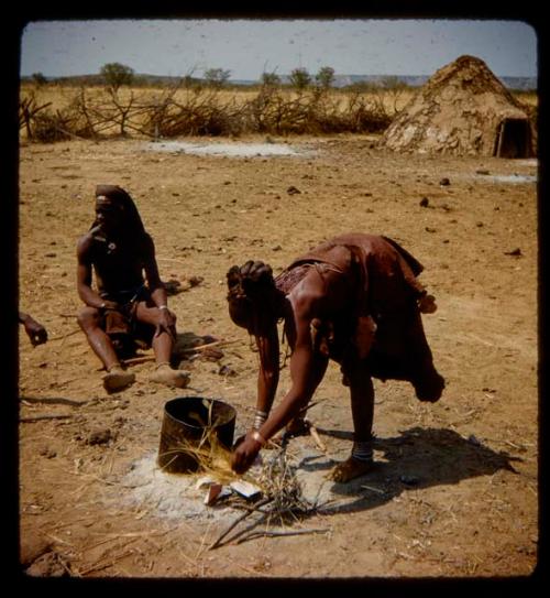 Woman putting grass on a fire, with a man sitting near her