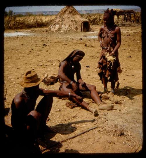 Woman standing, holding grass in her hand, with two men sitting near her