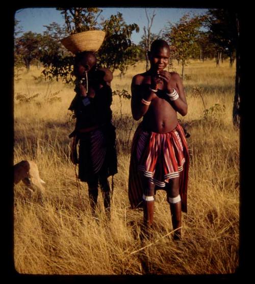 Two women standing, one carrying a basket on her head and a baby on her back