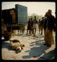 Group of people standing around a truck, with a goat and a dead bird on the ground next to it