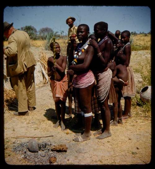 Women wearing bead skirts standing next to a boy holding reeds, with Merl La Voy and another man standing near them