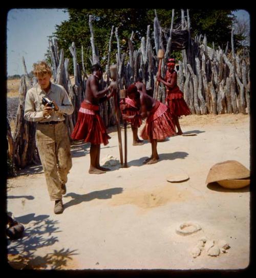 Four of Nehemiah's wives pounding something in holes in the ground with long poles, with John Marshall walking next to them carrying a film camera