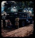 John Marshall kneeling behind an expedition truck, holding a Polaroid camera, with two men standing next to him